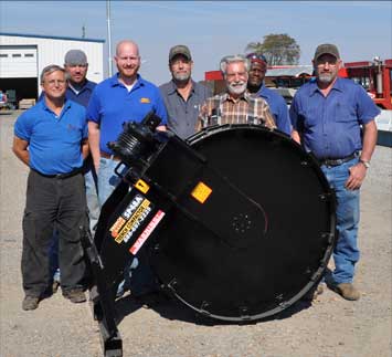 A group photo of the trench grader team standing next to a custom soil compaction wheel.